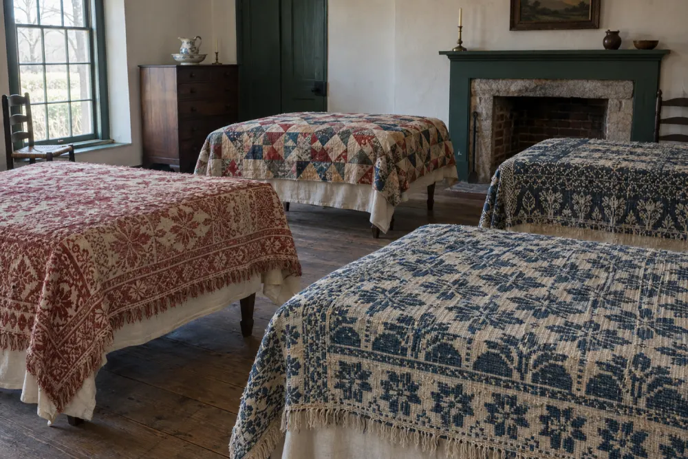 The upstairs bedroom set for the coverlet exhibition: three woven coverlets in madder red, indigo and undyed cotton and a patchwork quilt laid flat on low tables, plaster walls, a green-painted mantel, raking light from the window.